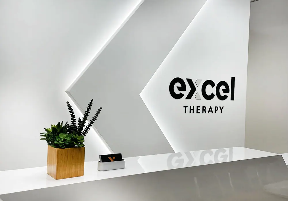 The front desk at Excel Therapy, in front of the lit white chevron wall with the Excel Therapy logo
